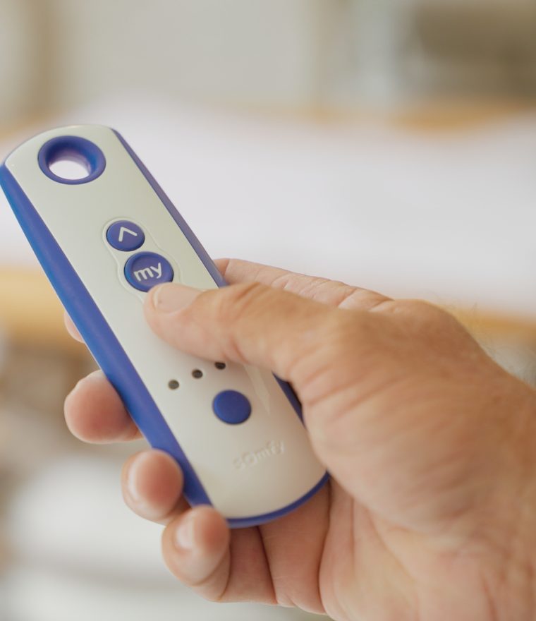 A hand holding a Somfy brand motorized screen remote control with a circular top button, directional buttons, and blue accents